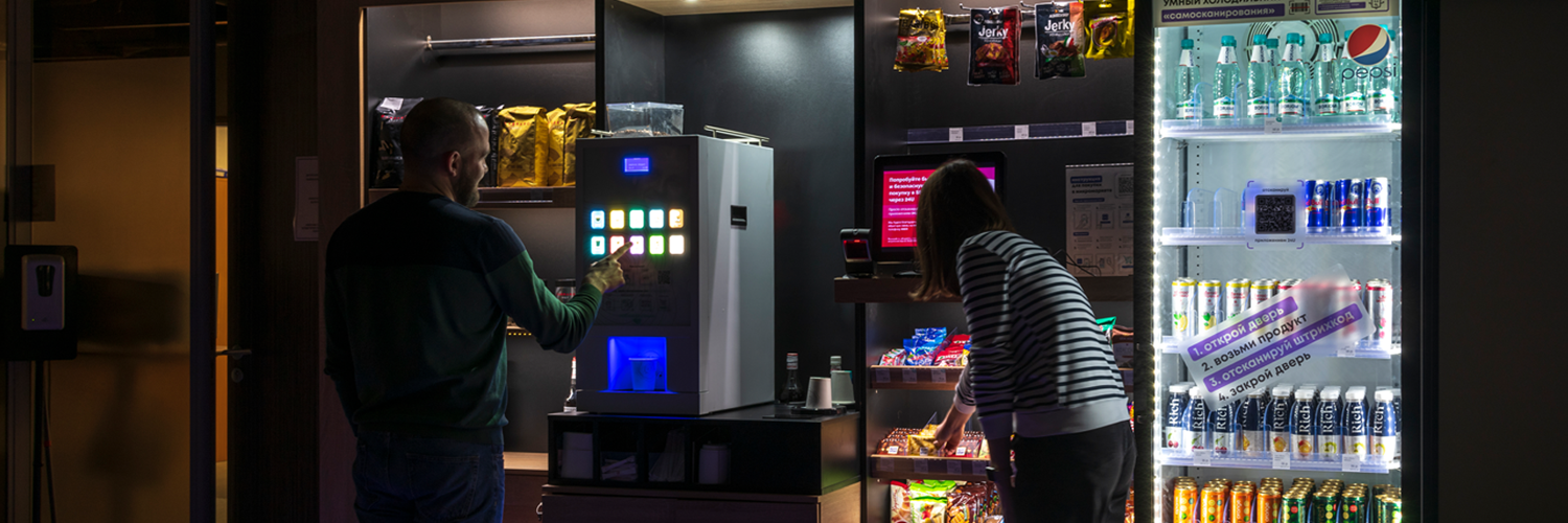 Two people using a modern micro market with a vending machine and refrigerated cooler stocked with drinks and snacks
