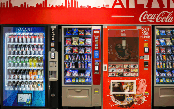 Vending machines in a public area serving Coca-Cola drinks, packaged snacks, and ice cream, powered by MONEXgroup cashless payment technology.