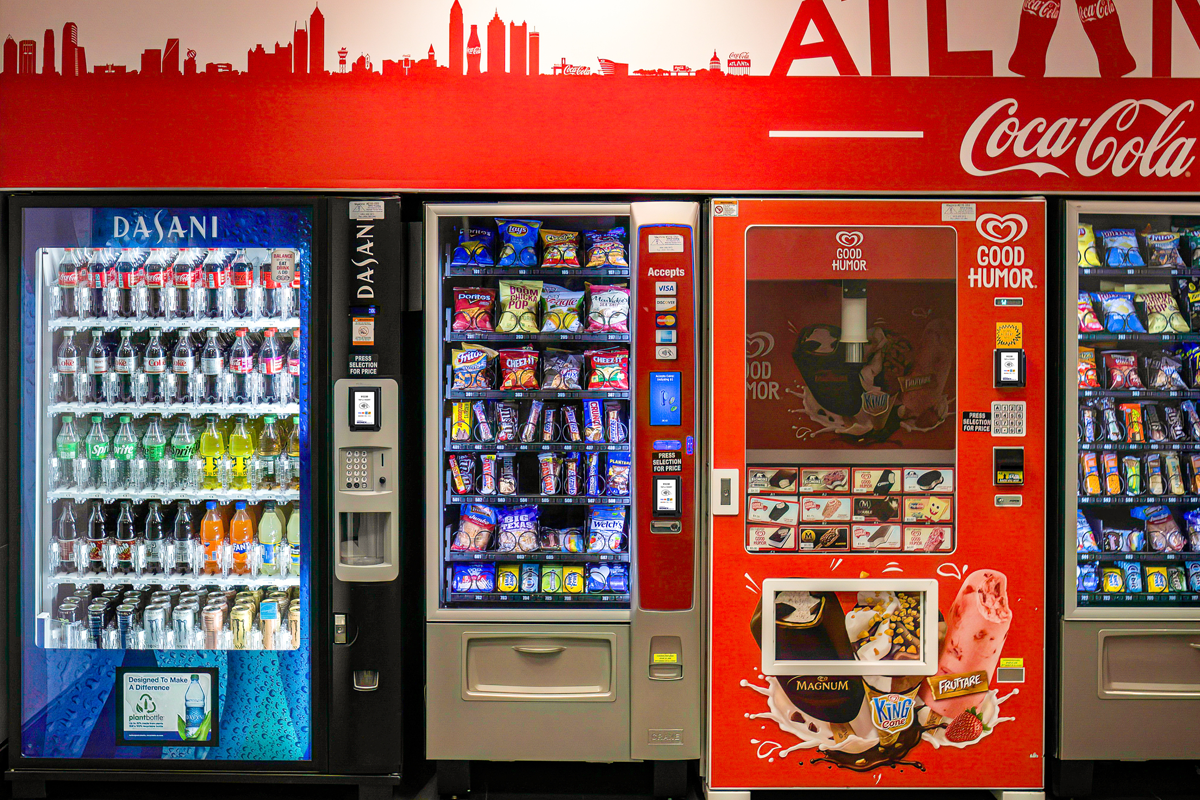Vending machines in a public area serving Coca-Cola drinks, packaged snacks, and ice cream, powered by MONEXgroup cashless payment technology.