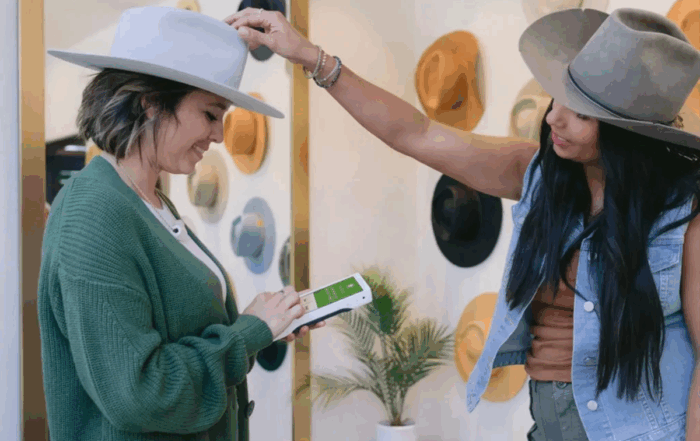 Retail employee using a mobile POS device while assisting a customer trying on hats in a boutique