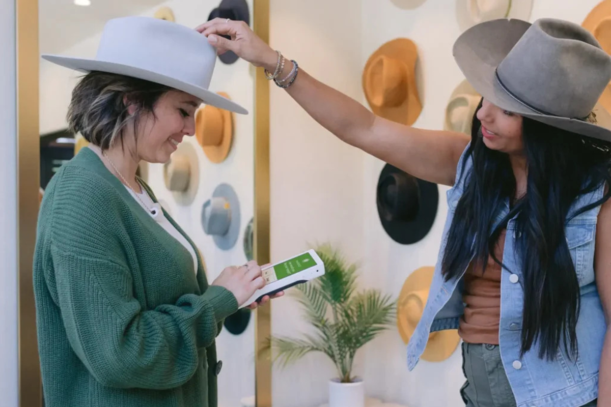 Retail employee using a mobile POS device while assisting a customer trying on hats in a boutique